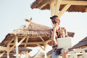 Portrait of a handsome young student guy working with a laptop and talking on the phone with his friends on a sunny summer day on the beach under a thatched canopy