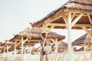 Portrait of a handsome young student guy working with a laptop and talking on the phone with his friends on a sunny summer day on the beach under a thatched canopy