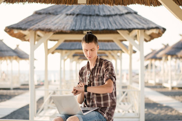 A young student guy looks at the time and wants to finish classes with a laptop in the fresh air under the thatched roof on a sunny summer day. Concept of a break in study and rest