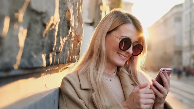 Portrait of woman in autumn clothes and sun glasses writing message on her smartphone outdoors. Girl using digital gadget, scrolls through social media on device, reading news on app, at sunset.