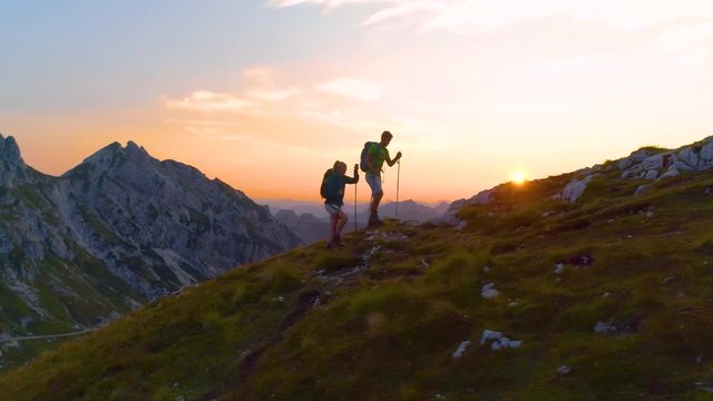 AERIAL, LENS FLARE: Active young tourists hiking up a grassy mountain in the Alps at sunset. Carefree hiker couple enjoying a relaxing evening trip in the breathtaking Julian Alps. Tourists on a trek.