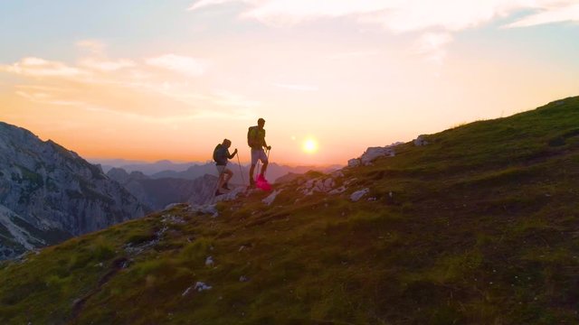 AERIAL, LENS FLARE: Carefree young trekkers on an active summer vacation hiking uphill on a picturesque summer morning. Tourist couple exploring the stunning Julian Alps at sunrise. Fun recreation.