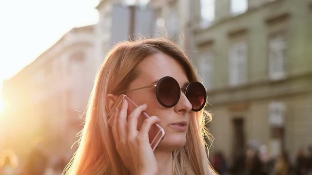 Close up portrait of beautiful trendy caucasian woman talking on smartphone, enjoying urban city lifestyle. Girl calling on mobile phone, wearing sun glasses and coat, outdoors.
