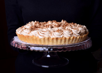Banoffee pie topped with whipped cream and sprinkled with grounded coffee on a glass stand in female hands