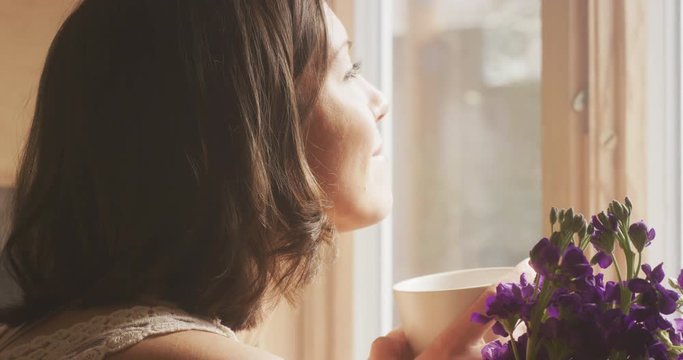 A sun filled window frame and purple flowers fill the frame.  A woman enters the frame with a mug of coffee.  She closes her eyes feeling the sun on her face.  She takes a sip from her mug and smiles.