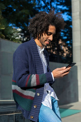 Young handsome African man using his smartphone with smile while sitting on a bench outdoors in sunny day