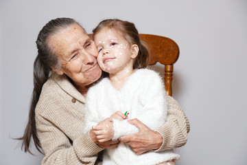 Сute gray long haired grandmother in knitted sweater hugs granddaughter with chicken pox, white...