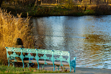 Alte himmelblaue gusseiserne Parkbank im herbstlichen Sonnenschein am Seeufer mit einer Person von...