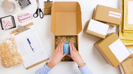 delivery, mail service, people and shipment concept - woman packing mug to parcel box with straw filler and crossing item off form on clipboard at post office - Powered by Adobe