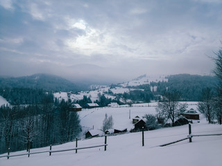 Winter countryside mountain landscape with wooden houses covered with snow, forests in the misty distant background. Picturesque and peaceful wintry scene. European resort location. Blue toning
