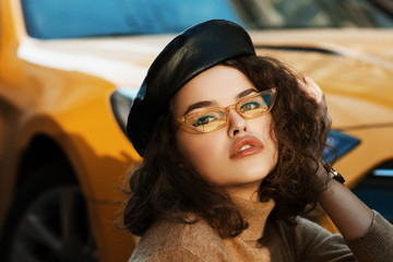 Outdoor close up fashion portrait of young beautiful confident woman wearing trendy orange sunglasses, stylish leather beret, cashmere turtleneck, posing in street, near the car. Copy, empty space 