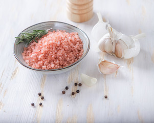 A sprig of rosemary lies in a white bowl with pink Himalayan salt and around other spices.