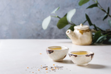 Hot green tea in two traditional chinese clay ceramic cup and teapot standing on white marble table. With green branch at background.