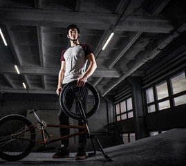 Full body portrait of a young man in protective helmet with his bike standing in a skatepark indoors