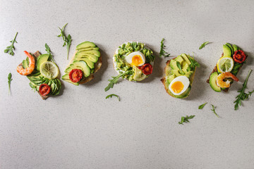 Variety of vegetarian sandwiches with sliced avocado, sun dried tomatoes, egg, shrimps, arugula served over white grey spotted background. Flat lay, space