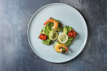 Vegetarian sandwich with sliced avocado, sun dried tomatoes, shrimps, lemon served on ceramic plate over blue texture background. Flat lay, space