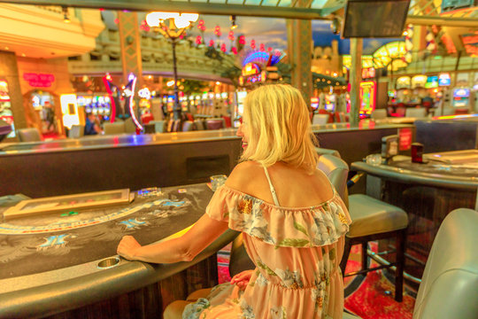 Back side of caucasian blonde woman gambling in casino with bright lights. Lifestyle girl waiting to play at blackjack in Las Vegas, Nevada, United States. Gambler addict concept