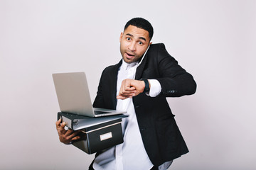 Busy hard-working guy in white shirt and black jacket talking on phone, holding folders, laptop, looks astonished at watch on white background. Office worker, modern technology, career