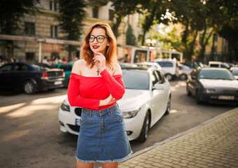 Fototapeta premium Attractive redhaired woman in eyeglasses, wear on red blouse and jeans skirt posing at street against white sport car.