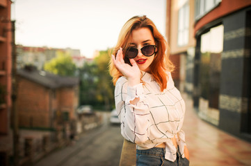 Fototapeta premium Attractive redhaired woman in sunglasses, wear on white blouse posing at street against modern building.