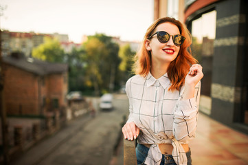 Attractive redhaired woman in sunglasses, wear on white blouse posing at street against modern building.
