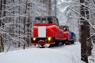 An old vintage red train with small blue carriages rides through the winter snowy forest. Fabulous winter landscape. Narrow gauge railway. Logistics railway transport