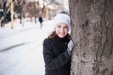 Young girl hugs a tree and smiles in the winter, having a rest in the city after school. Everywhere lies the snow