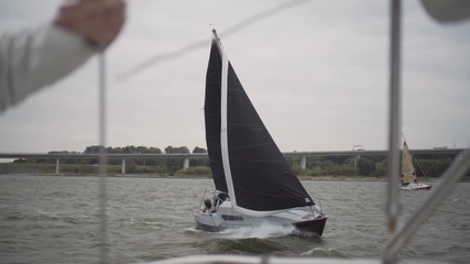 Sailing ship with black sails passes by on the ocean on a windy day with lots of waves and a bridge in the background.