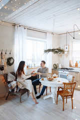 Fototapeta na wymiar Young caucasian couple sitting at home kitchen, hugging, smiling, drinking tea. Young lovers having good time. Man and woman communicate sitting at a table in the dining room.Winter decor.