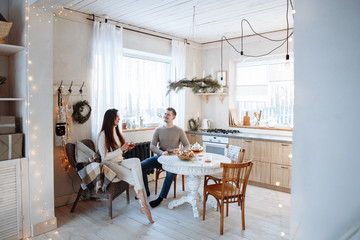 Young caucasian couple sitting at home kitchen, hugging, smiling, drinking tea. Young lovers having good time. Man and woman communicate sitting at a table in the dining room.Winter decor.