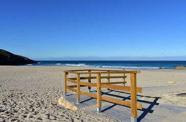Beach with wooden handrail and morning light. Golden sand and blue sea with waves and white foam. Sunny day, Galicia, Spain.
