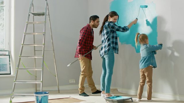 Beautiful Young Family are Showing How to Paint Walls to Their Adorable Small Daughter. They Paint with Rollers that are Covered in Light Blue Paint. Room Renovations at Home.