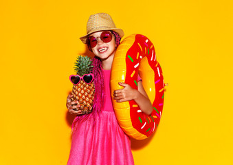 Funny happy child girl in summer pink dress with pineapple and swimming circle on yellow background.