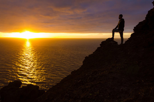 Hiker silhouette standing on cliff edge rock staring at sunset in Gran Canaria, Spain. Fearless man enjoys splendid twilight by the sea in Canary Islands. Visionary, adventure, challenge concepts