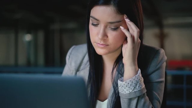 Portrait of a stressed, young brunette business woman holding her face whilst looking at her laptop in a modern office