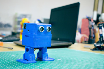 3D printed Funny dancing blue robot on the background of devices and laptop. Robot model printed on automatic three dimensional 3d printer. Additive manufacturing and robotic automation technology.