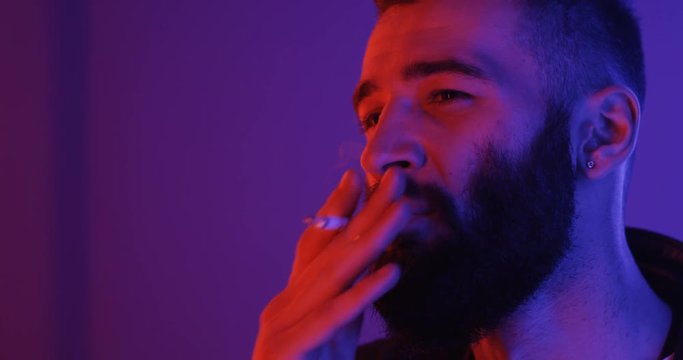 Close up of the handsome bad young guy with a beard smoking a cigarette in the neon pink and blue light and then smiling to the camera. Portrait.