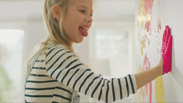Happy Little Girl with Hands Dipped in Yellow and Pink Paint Puts a Colorful Handprint on the Wall. She is Having Fun and Laughs. Home is Being Renovated.