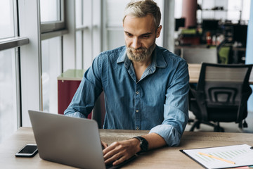 The working process. Stylish guy in a blue shirt with a beard working on a laptop in his office