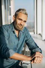 Fototapeta premium Portrait of a handsome stylish brutal man with a beard and blue shirt by the window