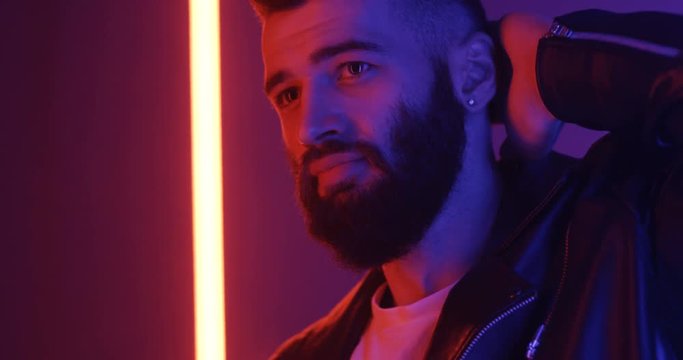 Close up of the attractive cool young bad guy with a black beard looking at the side and straight in the neon light.