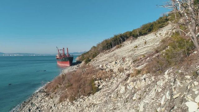 View on big red cargo ship in blue sea near coastline with people against clear blue sky. Shot. Marine landscapes
