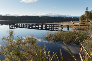 The wooden pier in Lake Mahinapua reflected on a calm, sunny morning. Ruatapu, West Coast New Zealand.