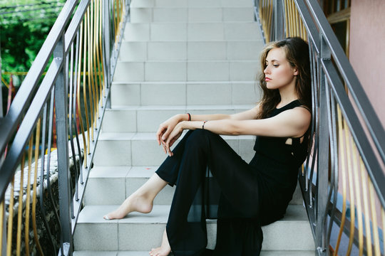 Portrait of sad glamour woman with long beautiful hair seating outdoor on stairs wearing black dress