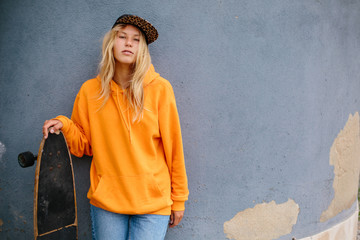 City portrait of positive young female wearing orange hoody and baseball cap holding skateboard. Grey blue wall on background © Annatamila