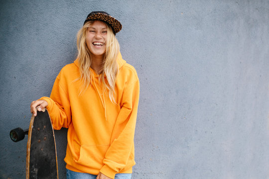 City portrait of positive young female wearing orange hoody and baseball cap holding skateboard. Grey blue wall on background