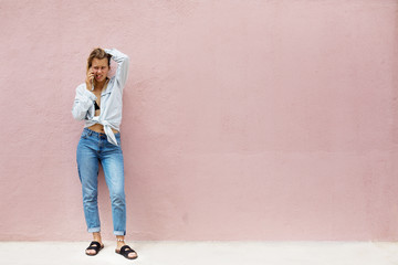 Young sad girl talking by mobile phone, standing outdoor with pink wall on background. Space for text