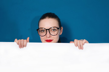 Young woman wearing glasses keeping blank paper. Blue background, studio portrait