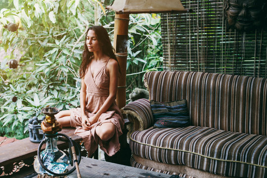 Fashion woman wearing natural fabrics dress seating in meditation pose outdoor on  striped couch in jungle forest resort