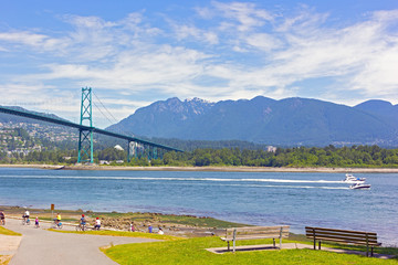 A trail in Stanley Park with locals and tourists on a beautiful day in summer, Vancouver BC, Canada. Landscape with Lions Gate Bridge, motorboats and mountainous ridge with snowy peaks.